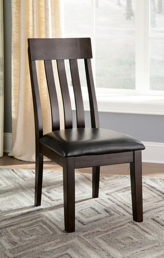 A Haddigan Dining UPH Side Chair (2/CN) with a dark wood frame and black faux leather upholstered seat sits on a patterned rug by a window with beige curtains.