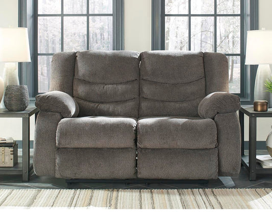 The Tulen Reclining Loveseat in gray chenille sits between two side tables with lamps, positioned in front of large black-framed windows.