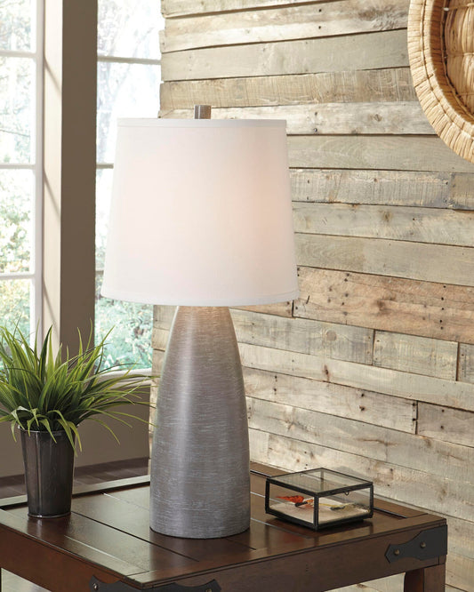 The Shavontae Poly Table Lamp (2/CN) with a white shade sits on a wooden table beside a potted plant and glass box, set against a wood-paneled wall.