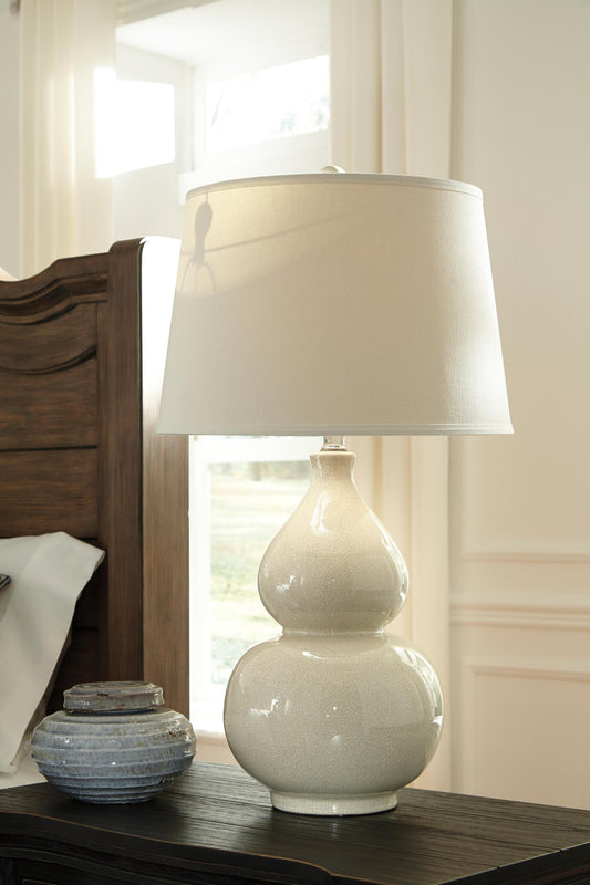 The Saffi Ceramic Table Lamp (1/CN) with a fabric shade sits on a dark wood nightstand by a wooden headboard and decorative jar, providing stylish contemporary lighting.