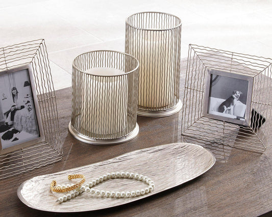 The Dympna Accessory Set (5/CN), featuring metal photo frames, hurricane candle holders with pillar candles, a pearl necklace, and a gold bracelet, is arranged on a wooden surface with a decorative tray.