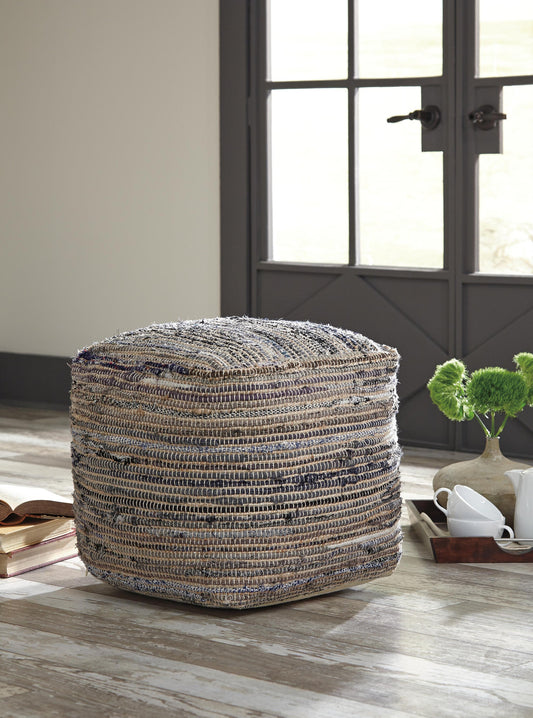 The Absalom Pouf sits hand-stitched on a wooden floor in a room with a gray door, books, cups on a tray, and a small vase with green flowers.