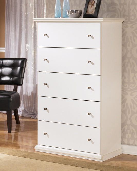 The Bostwick Shoals Five Drawer Chest, in a white cottage-chic style, sits on a wooden floor for stylish clothing storage. On top are vases, a bowl, and a framed photo; a black chair is placed to the left.