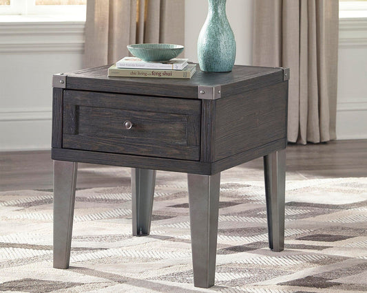 The Todoe Rectangular End Table features a dark wood drawer, metal legs, and corner accents. Topped with a vase, bowl, and books on a patterned rug in a bright room, it stylishly blends rustic-industrial charm with function.