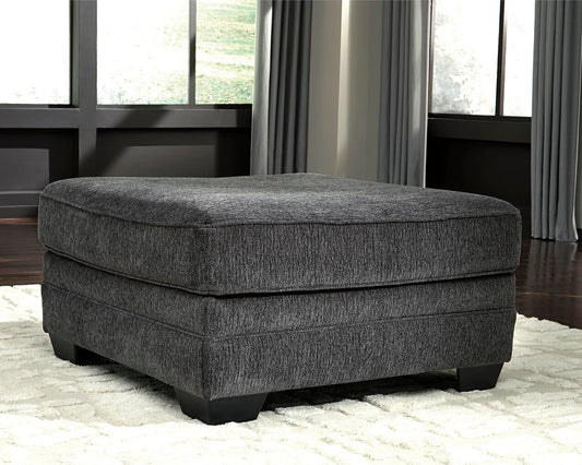 The Tracling Oversized Accent Ottoman in slate gray fabric sits on a white rug in a living room with large windows and gray curtains, enhancing the contemporary style.