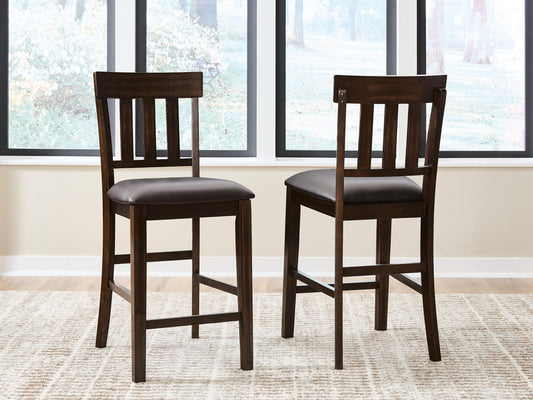 Two Haddigan Upholstered Barstools with slat backs and faux leather seats are placed on a light patterned rug in front of large windows.