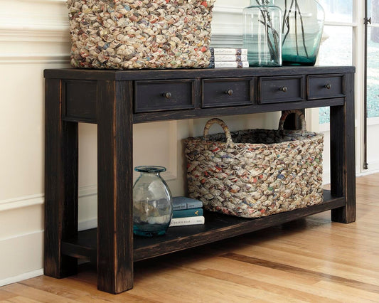 The Gavelston Sofa Table has a weathered black finish, four small drawers, and is styled with woven baskets, glass vases, and books on and below it. It sits against a white wall on a wooden floor.