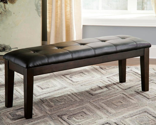 The Haddigan Large UPH Dining Room Bench, featuring a dark brown tufted faux leather seat, is set on a patterned rug in a bright room with light curtains.