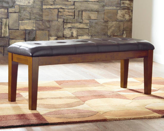 The Ralene Large UPH Dining Room Bench with a dark cushioned seat sits on a geometric-patterned rug in front of a stone accent wall.