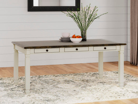 The Bolanburg Rectangular Dining Room Table with white legs and a dark top sits on a light patterned rug, decorated with a vase of branches, bowls, and a plate of fruit.