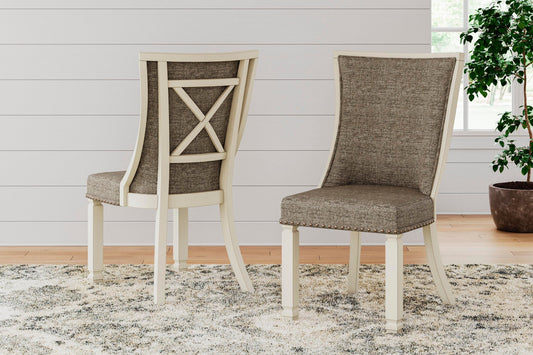 Two Bolanburg Dining UPH Side Chairs with cushioned seats and light wood frames in an antiqued white finish sit on a patterned rug in a bright room with white shiplap walls and a potted plant.
