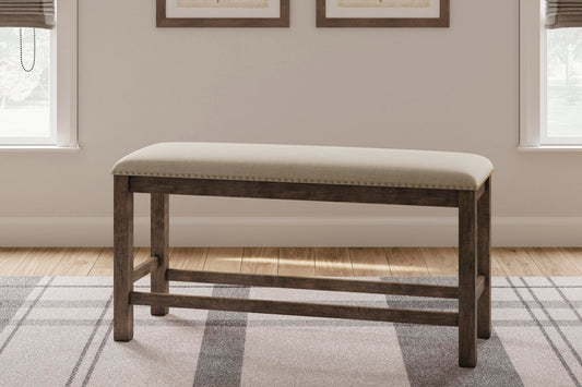 The Moriville Double UPH Bench, featuring a counter height and distressed nutmeg finish with a beige seat, sits on a patterned rug between two windows in a light-colored room.