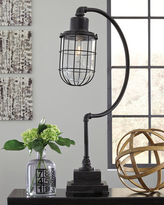 The Jae Metal Desk Lamp (1/CN), in black with a curved arm, sits on a table beside a glass vase of green flowers and a decorative metal sphere, with a window in the background.