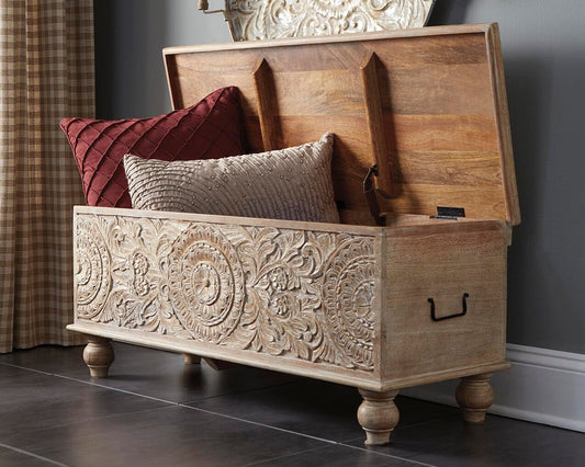 The Fossil Ridge Storage Bench in antique white features hand-carved floral patterns, an open lid revealing two decorative throw pillows inside, and is placed on a tiled floor near a window.