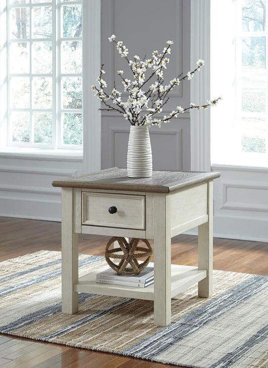 The Bolanburg Rectangular End Table, with its two-tone distressed finish, features a smooth-gliding drawer and shelf. It sits on a striped rug and is topped with a white vase of flowers in a bright, sunlit room.