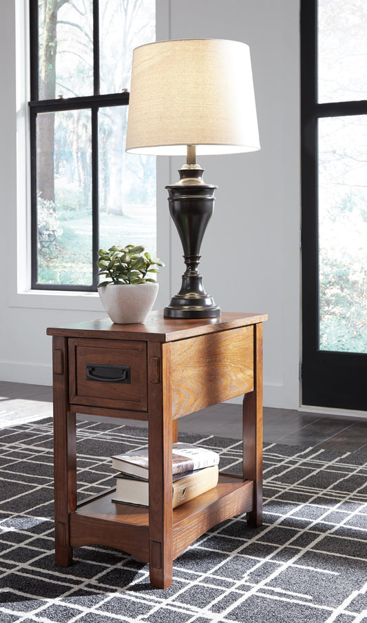 The Breegin Chair Side End Table, featuring a drawer, displays a black lamp, a small potted plant, and stacked books on its lower shelf. This stylish piece rests on a geometric-patterned rug by the window.