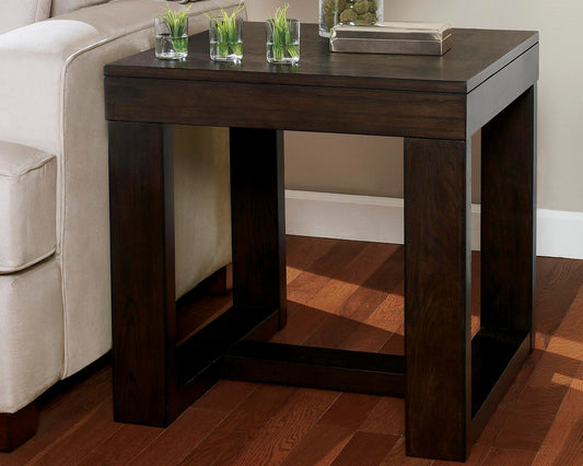 The Watson Square End Table, featuring a dark wood and modern design, stands by a beige sofa on a hardwood floor, finished with small plants and a book on top.