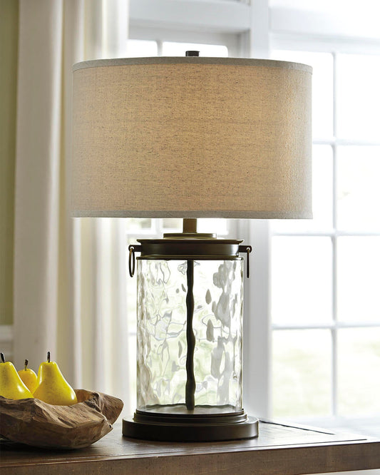 The Tailynn Glass Table Lamp, featuring a textured cylindrical base and beige fabric shade, sits on a wooden surface near a window beside a wooden bowl filled with yellow pears.