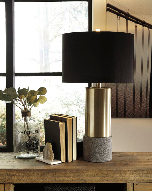 The Jacek Metal Table Lamp with a black shade sits on a wooden table alongside a glass vase with greenery and three books, placed in front of a window and framed wall art.