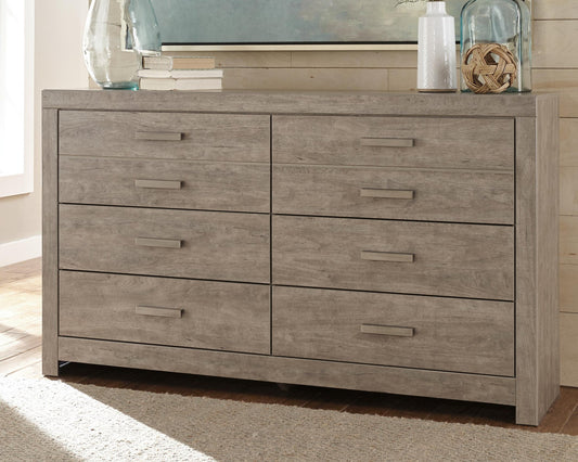 The Culverbach Six Drawer Dresser, featuring a gray weathered driftwood finish, is decorated with books, glass vases, and a rope ornament and sits on a light-colored floor.