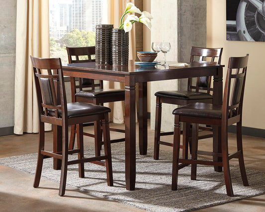 The Bennox DRM Counter Table Set (5/CN), featuring a wooden counter-height table and four matching high-back chairs, sits on a gray rug adorned with decorative items, bowls, and wine glasses near bright windows.