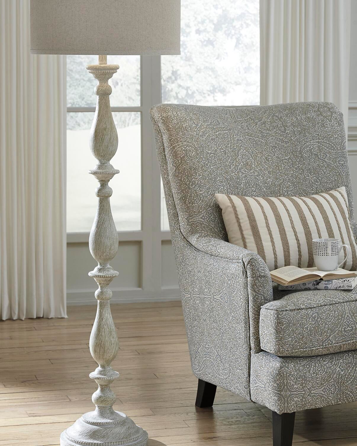 A patterned armchair with a striped cushion and cup sits beside the Bernadate Poly Floor Lamp, which features a classic candlestick design, in a bright room with wooden floors and large windows.