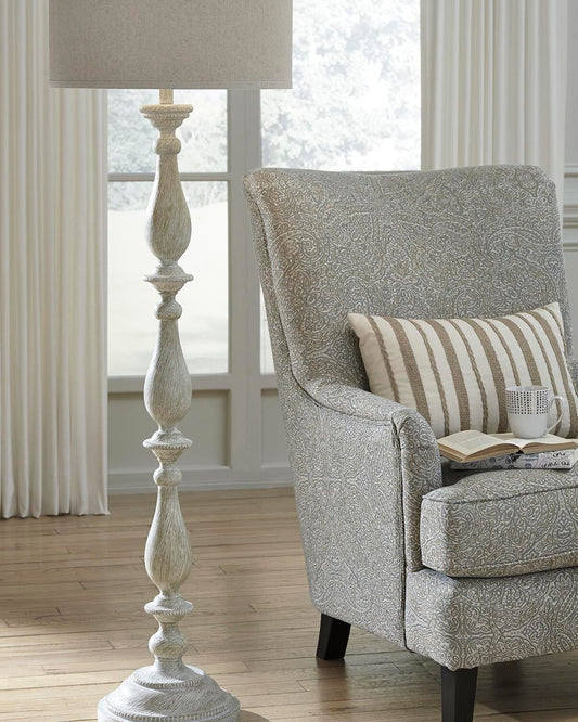 A patterned armchair with a striped cushion and cup sits beside the Bernadate Poly Floor Lamp, which features a classic candlestick design, in a bright room with wooden floors and large windows.