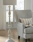 A patterned armchair with a striped cushion and cup sits beside the Bernadate Poly Floor Lamp, which features a classic candlestick design, in a bright room with wooden floors and large windows.