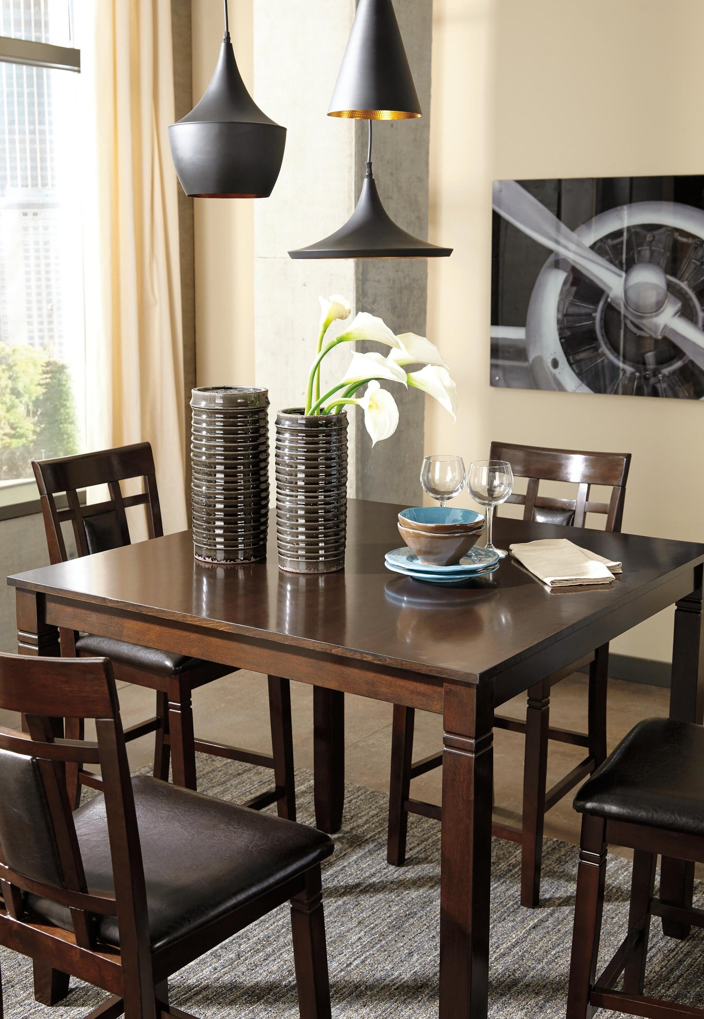 The Bennox DRM Counter Table Set (5/CN) features a dark wood counter-height table with four chairs, topped with plates, bowls, glasses, and a vase of white lilies. Three black pendant lights hang above and an aviation photo decorates the wall.