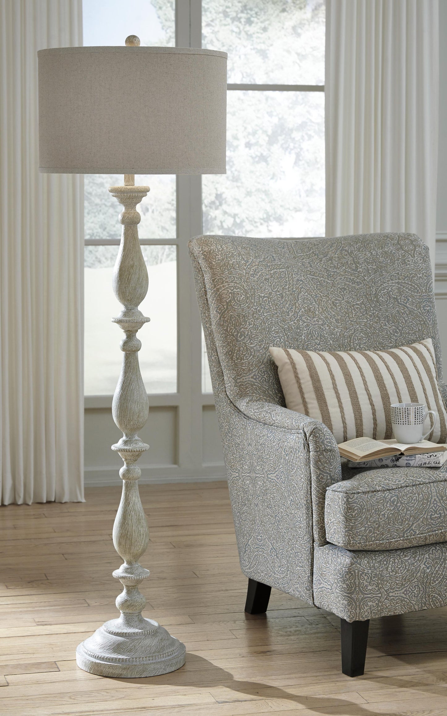 The Bernadate Poly Floor Lamp, featuring a candlestick design and whitewash finish, stands beside a patterned armchair with a striped pillow and book in a sunlit room with large windows and sheer curtains.