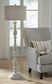 The Bernadate Poly Floor Lamp, featuring a candlestick design and whitewash finish, stands beside a patterned armchair with a striped pillow and book in a sunlit room with large windows and sheer curtains.