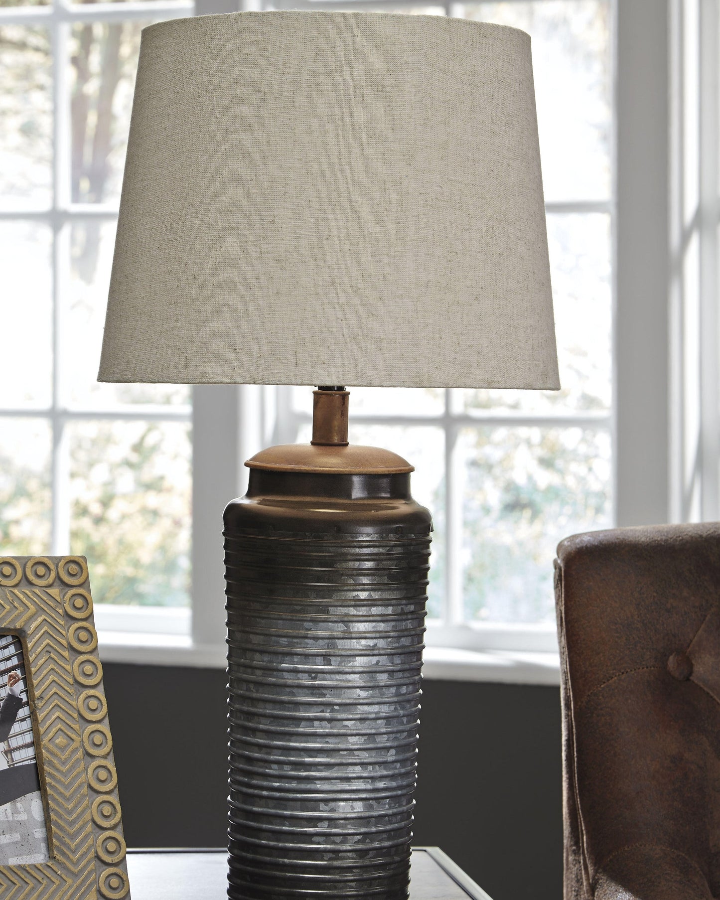 The Norbert Metal Table Lamp (2/CN), featuring a textured dark base and light fabric shade, is placed on a side table beside a decorative photo frame, with an armchair and window in the background.