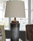The Norbert Metal Table Lamp (2/CN), featuring a textured dark base and light fabric shade, is placed on a side table beside a decorative photo frame, with an armchair and window in the background.