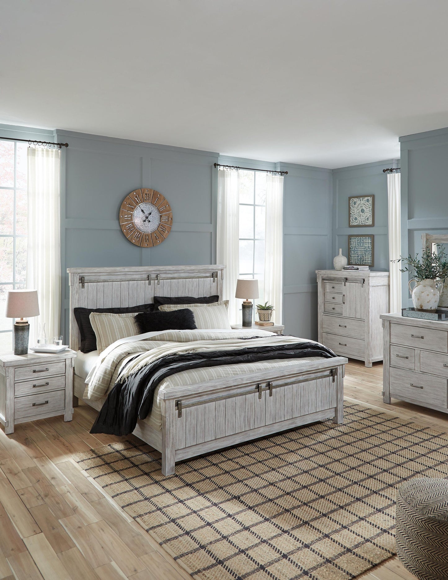A modern bedroom with light wood furniture, a double bed, two nightstands topped with Norbert Metal Table Lamps (set of 2), dressers, light blue walls, large windows with white curtains, and a patterned rug on a wooden floor.
