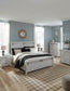 A modern bedroom with light wood furniture, a double bed, two nightstands topped with Norbert Metal Table Lamps (set of 2), dressers, light blue walls, large windows with white curtains, and a patterned rug on a wooden floor.