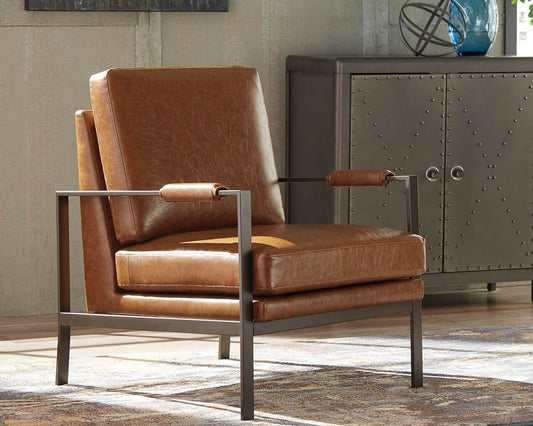 The Peacemaker Accent Chair in brown faux leather with a metal frame sits on a patterned rug in a modern room, beside a gray cabinet featuring rivet details.