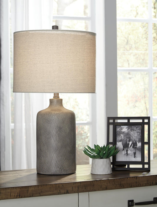The Linus Ceramic Table Lamp, a small potted plant, and a framed photo rest on a wooden table in front of large windows with sunlight streaming in.