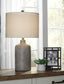 The Linus Ceramic Table Lamp, a small potted plant, and a framed photo rest on a wooden table in front of large windows with sunlight streaming in.