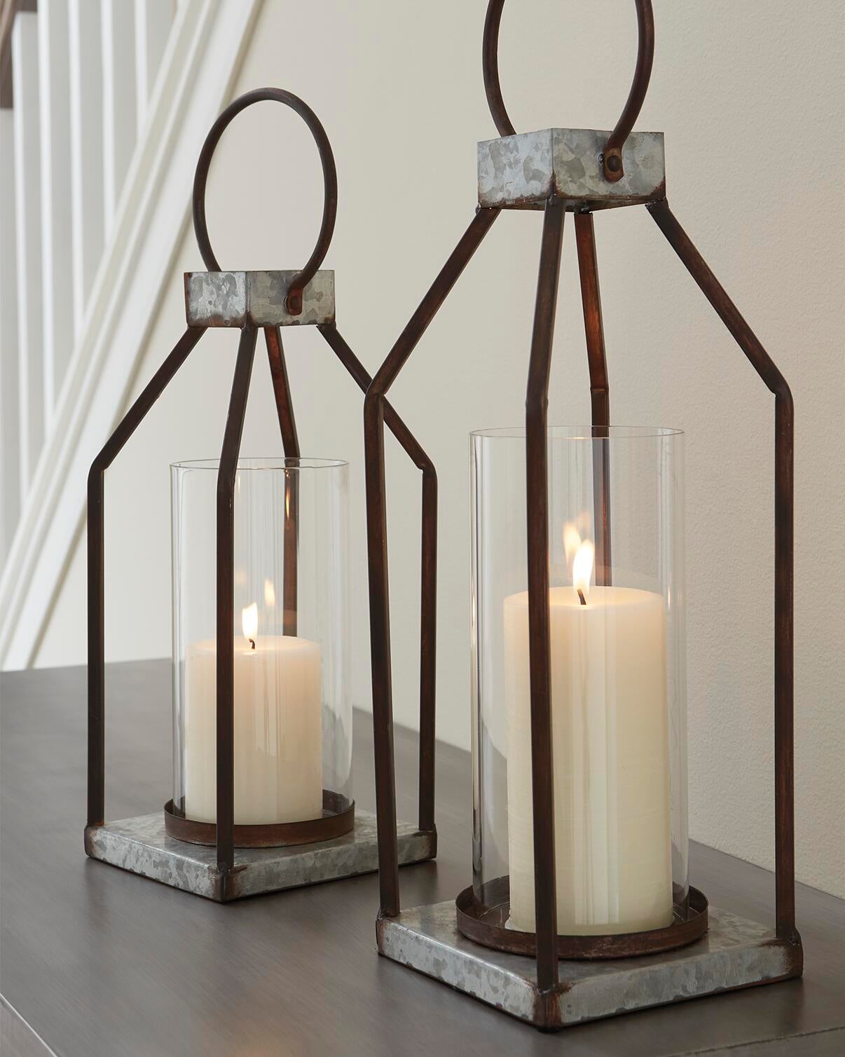 The Diedrick Lantern Set (2/CN), featuring glass enclosures and white pillar candles, casts a warm glow on a wooden surface beside the staircase—an elegant indoor-outdoor candle holder lantern duo.