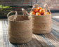 The Brayton Basket Set (2/CN) sits on a striped rug—one basket is empty, the other filled with oranges—offering a stylish solution for home organization.