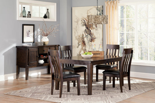 A dining room featuring the Haddigan RECT Dining Room EXT Table seating up to 8, six chairs on a patterned rug, a sideboard with decor, wall art, and large windows dressed in light curtains.