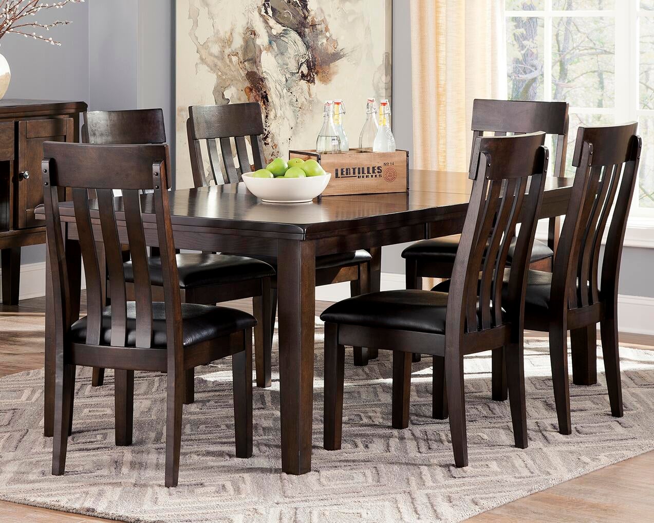 The Haddigan RECT Dining Room EXT Table, shown with six matching chairs, a bowl of green apples, and glass bottles atop a patterned rug in a bright room with large windows, features a butterfly leaf extension to seat up to 8.