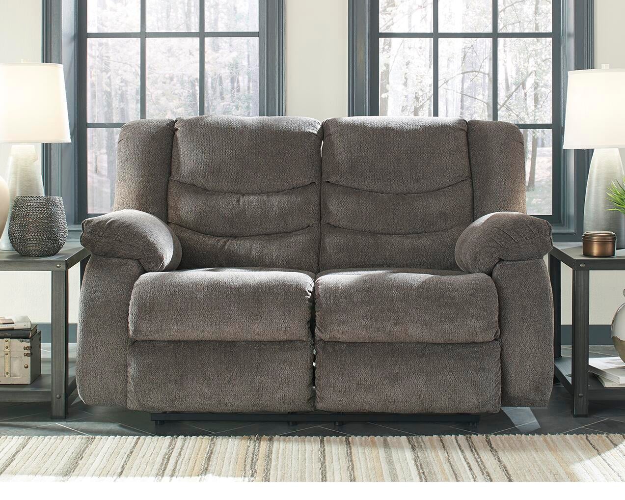 The Tulen Reclining Loveseat in gray chenille sits between two side tables with lamps, positioned in front of large black-framed windows.