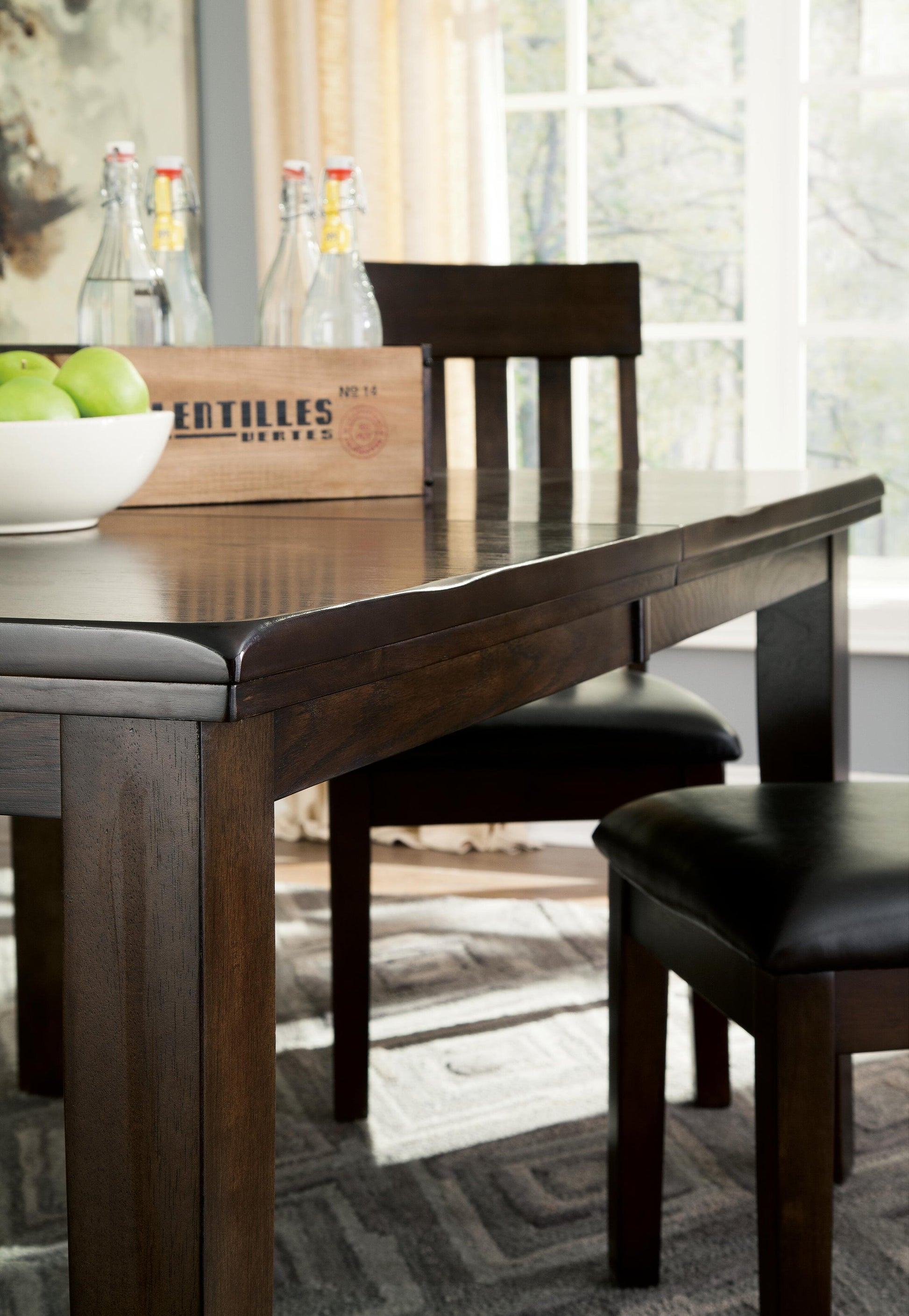 The Haddigan RECT Dining Room EXT Table, a dark wood farmhouse style with butterfly leaf extension, comes with two matching chairs. It’s shown in a sunlit room, topped with green apples and glass bottles in a wooden crate.