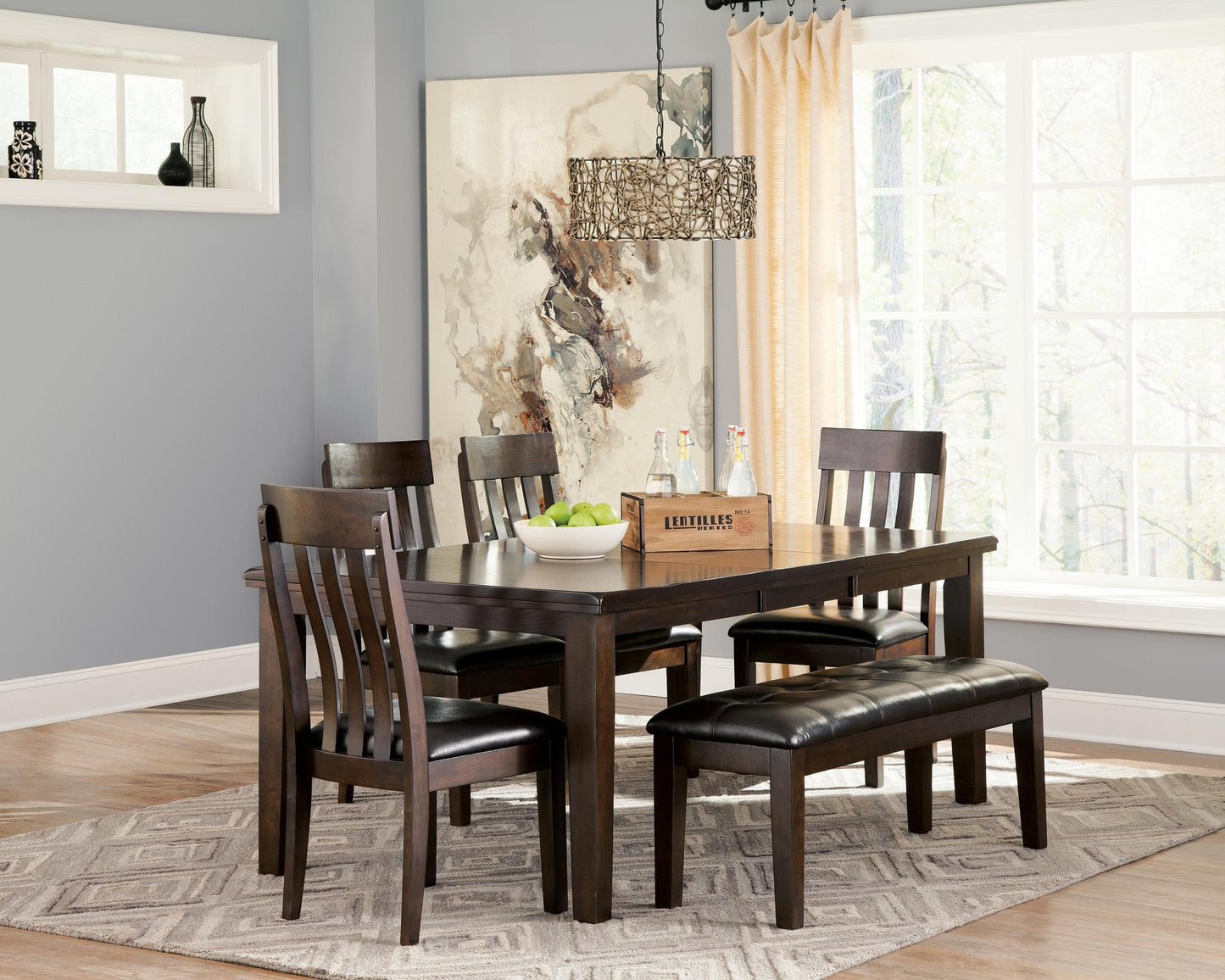 A dining room with a Haddigan RECT Dining Room EXT Table featuring a butterfly leaf, four matching chairs, a bench, a fruit bowl, drinks tray, abstract wall art, and a large window with light curtains.