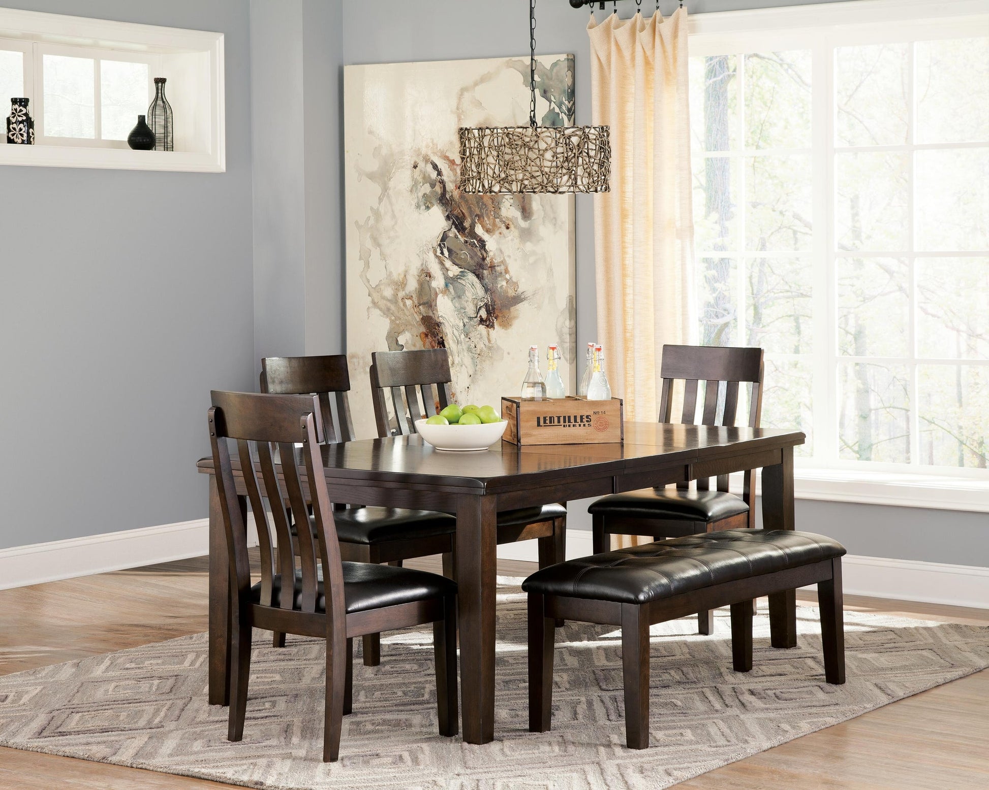 A dining room with a Haddigan RECT Dining Room EXT Table featuring a butterfly leaf, four matching chairs, a bench, a fruit bowl, drinks tray, abstract wall art, and a large window with light curtains.