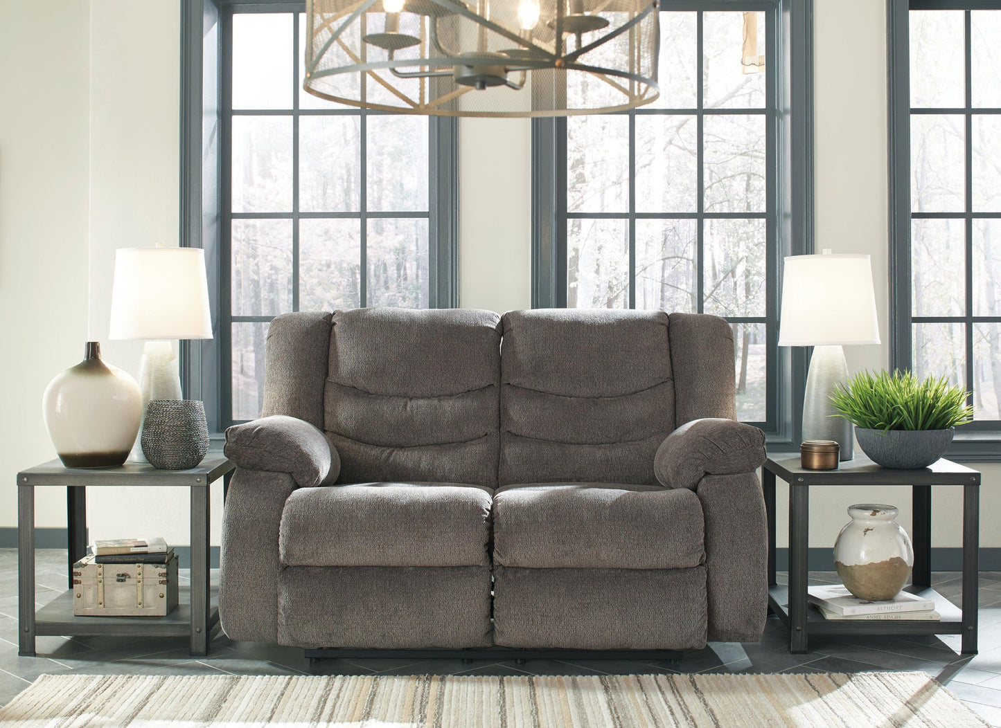 The Tulen Reclining Loveseat in soft gray chenille sits between two side tables with lamps and vases in a bright room featuring large windows and a geometric chandelier.