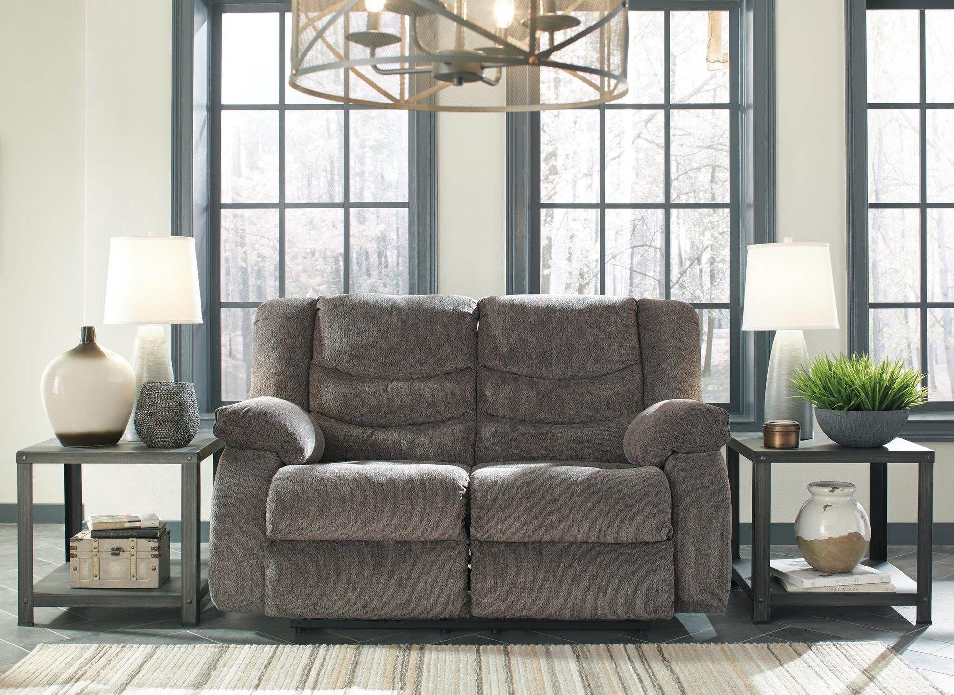 The Tulen Reclining Loveseat in soft gray chenille sits between two side tables with lamps and vases in a bright room featuring large windows and a geometric chandelier.