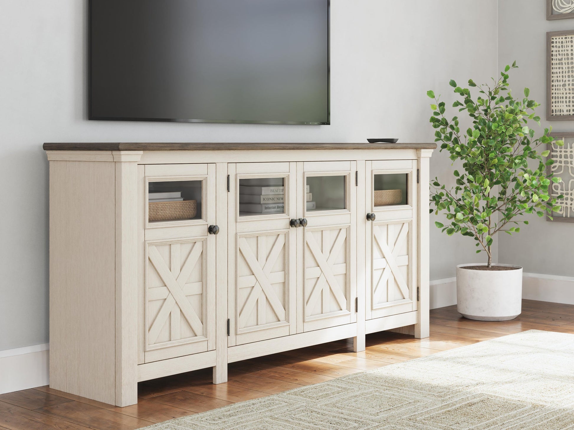 The Bolanburg Extra Large TV Stand, with glass and paneled cabinet doors in light wood, sits beneath a wall-mounted TV in a living room decorated with a potted plant and neutral accents.