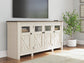The Bolanburg Extra Large TV Stand, with glass and paneled cabinet doors in light wood, sits beneath a wall-mounted TV in a living room decorated with a potted plant and neutral accents.